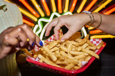two hands reaching for fries in a red basket with red checkered parchment in front of a yellow and orange neon sign
