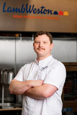 A chef in a white uniform stands with arms crossed in a commercial kitchen. 