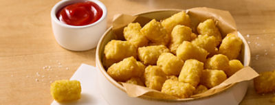 A bowl of golden, crispy tater tots is served on a wooden surface. The tots sit in a round dish lined with brown parchment paper. A small cup of red ketchup is placed beside the bowl for dipping. Coarse salt crystals are scattered around, adding to the appetizing presentation.