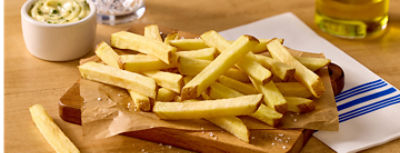 A pile of golden French fries is arranged on parchment paper over a wooden serving board. Coarse salt is sprinkled around the fries, emphasizing their crisp texture. In the background, a small bowl of creamy dip, a glass container of oil, and a striped napkin suggest a casual dining or snack setting. The warm lighting highlights the appetizing color and simple presentation of the fries.