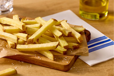 A pile of golden French fries is arranged on parchment paper over a wooden serving board. Coarse salt is sprinkled around the fries, emphasizing their crisp texture. In the background, a small bowl of creamy dip, a glass container of oil, and a striped napkin suggest a casual dining or snack setting. The warm lighting highlights the appetizing color and simple presentation of the fries.
